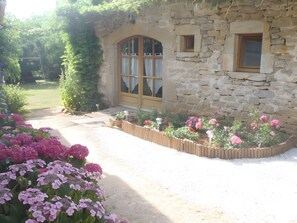 Ein Steinhäuschen mit einer Holztür und Fenstern, umgeben von einem Garten mit rosa Blumen.