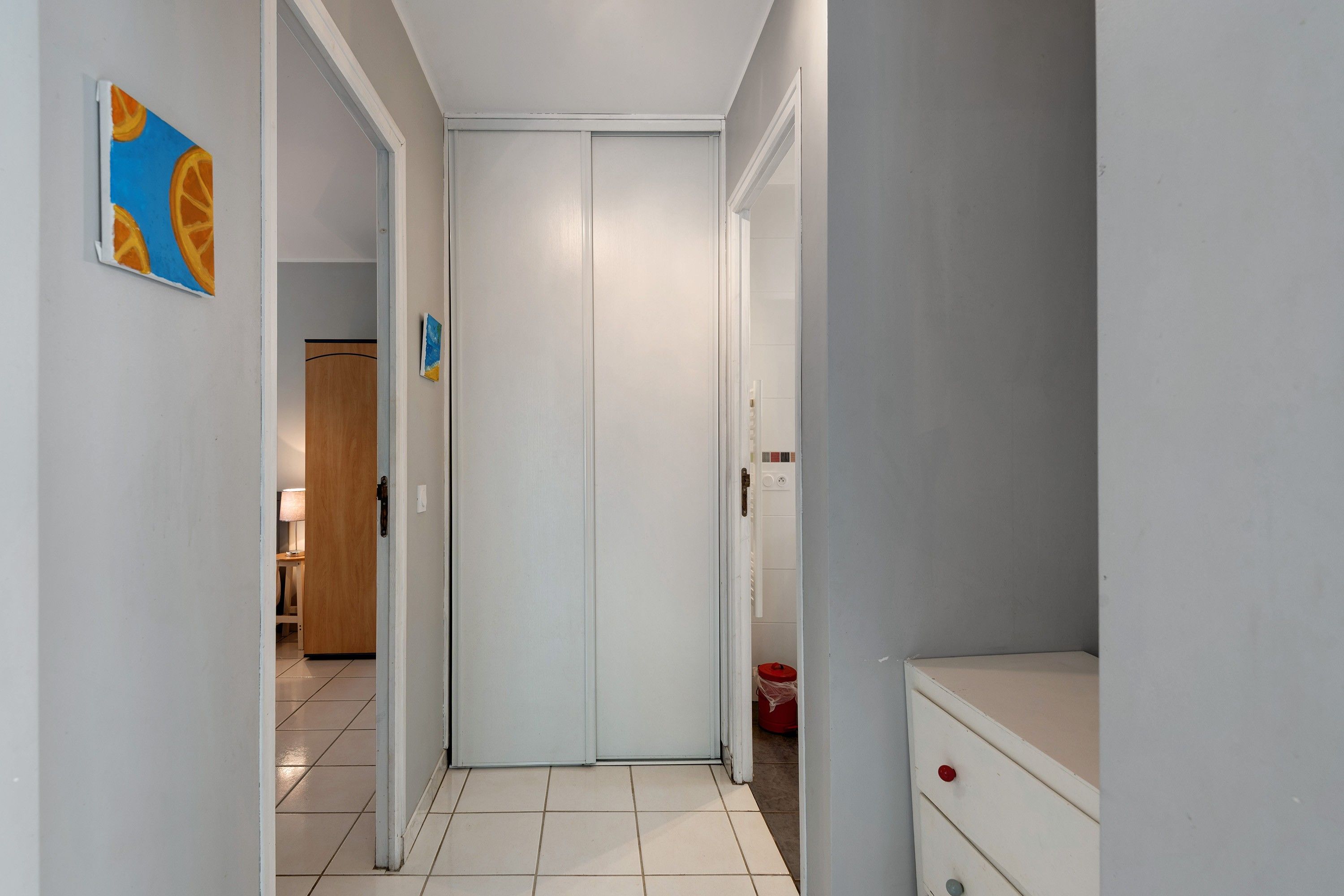 Un couloir étroit, avec une porte blanche au fond, une porte en bois sur la gauche et un seau rouge sur la droite.