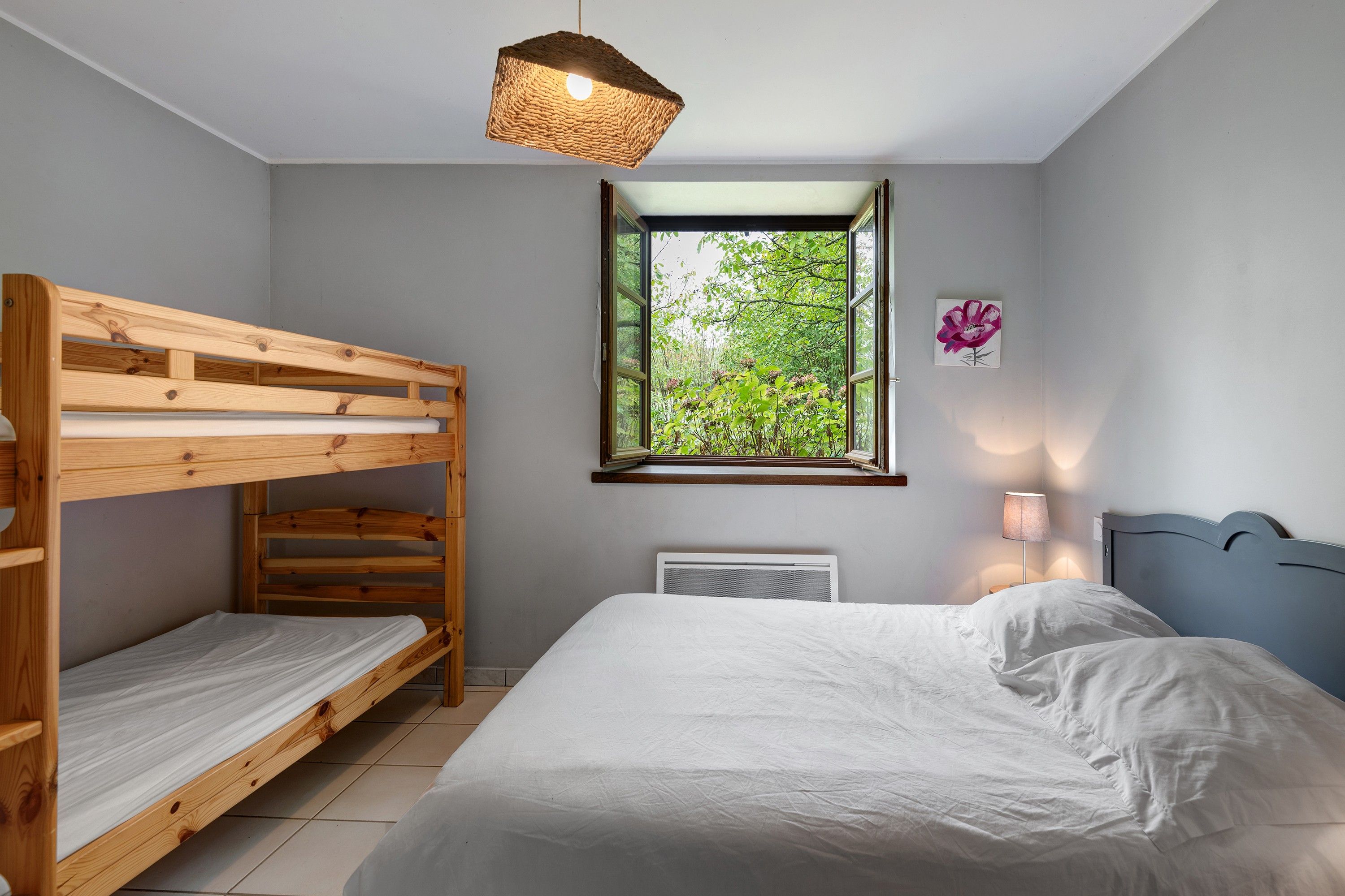A room with a bunk bed, a window with a view of greenery, a radiator, and a wall-mounted lamp.