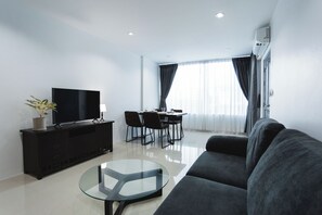 A modern living room with a flat-screen TV, a glass coffee table, a sofa, a dining area with chairs, and a large window with curtains. - The One Apartment (rawai)