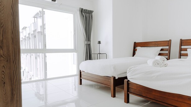 A hotel room with two wooden beds, a large window with curtains, and a view of buildings outside. - The One Apartment (rawai)