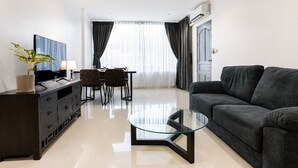 A modern living room with a glass coffee table, dark sofa, and a dining area with chairs and a TV. - The One Apartment (rawai)