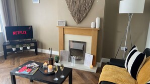 A living room with a fireplace, a television displaying 'NETFLIX,' a coffee table with magazines and candles, and a sofa with a throw blanket.