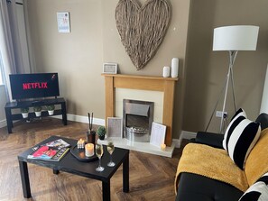 Un salón con chimenea, un televisor mostrando 'NETFLIX', una mesa de centro con revistas y velas, y un sofá con una manta.