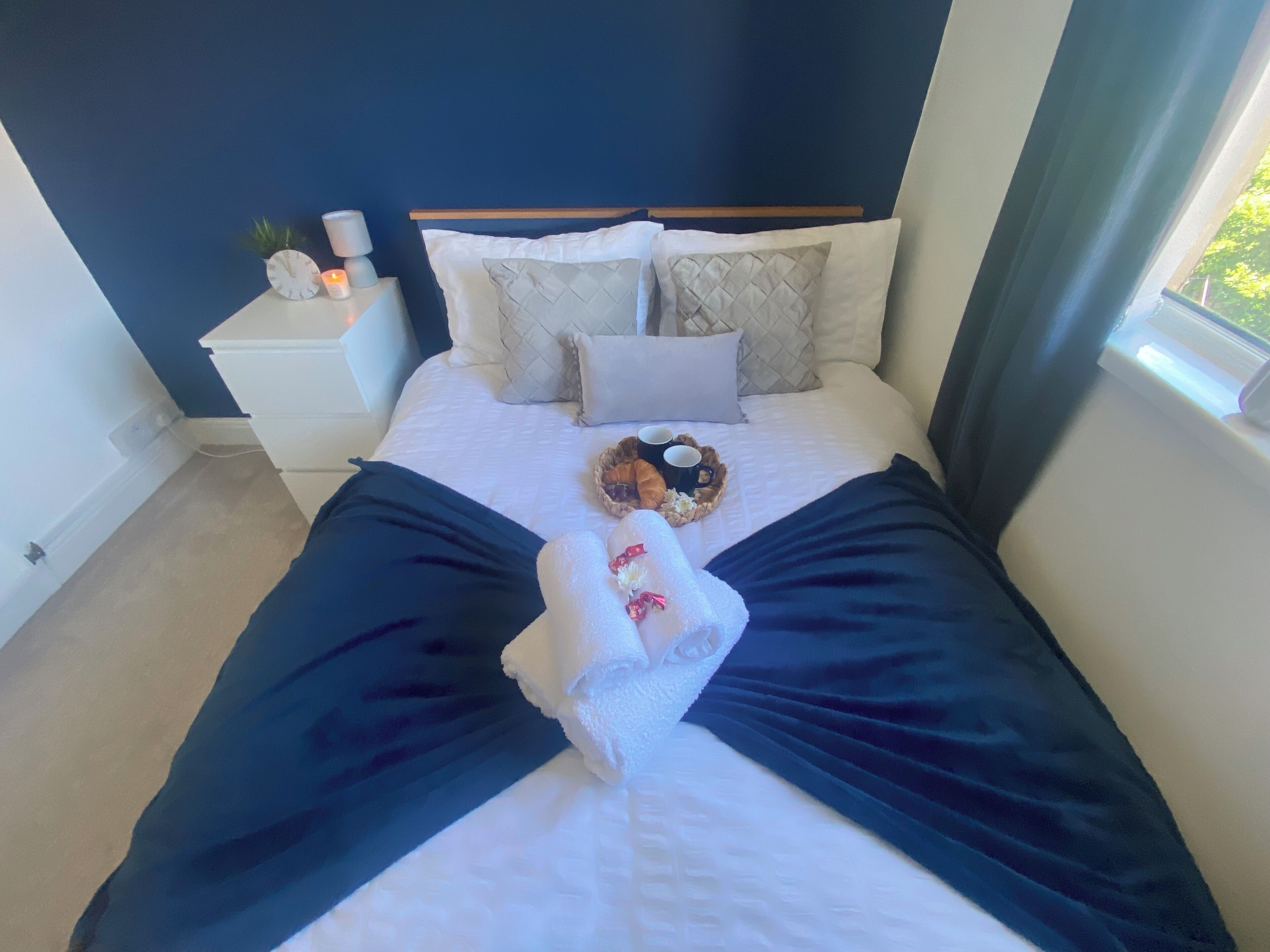 Un lit bien fait, recouvert d’une couvre-lit bleu foncé, avec deux oreillers, une table de chevet sur laquelle se trouvent une horloge et une bougie, et un panier contenant de la nourriture et des boissons.