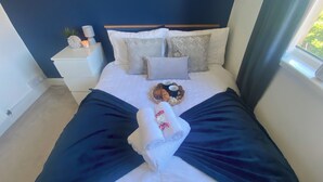 A neatly made bed with a dark blue bedspread, two pillows, a bedside table with a clock and a candle, and a basket with food and drinks.