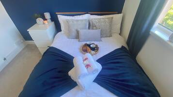 Un lit bien fait, recouvert d’une couvre-lit bleu foncé, avec deux oreillers, une table de chevet sur laquelle se trouvent une horloge et une bougie, et un panier contenant de la nourriture et des boissons.