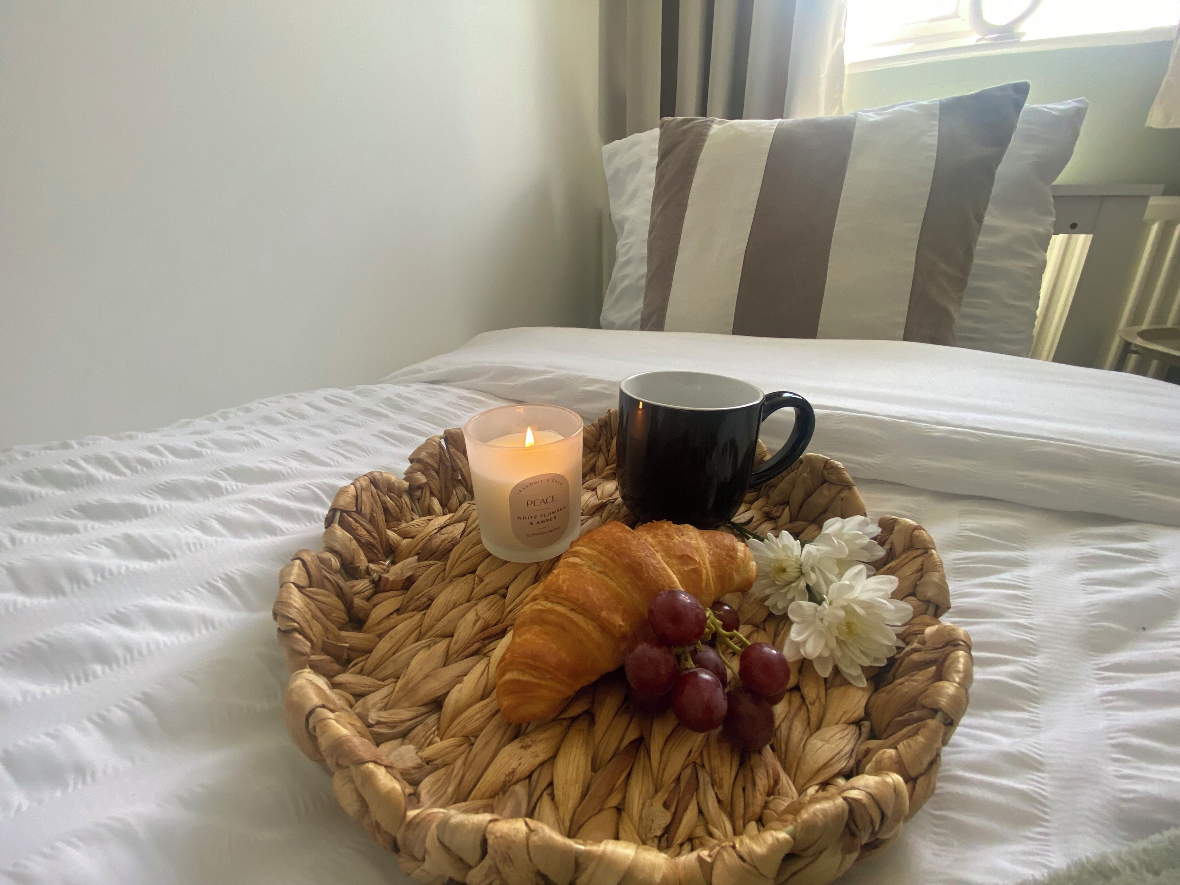 Ein Bett mit weißer Decke, ein gewebter Tablett mit einer Kerze, ein Croissant, Trauben und eine weiße Blume.