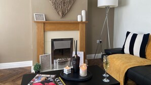 A living room with a fireplace, a black coffee table with magazines, a candle, a glass of wine, and a heart-shaped wall decoration.