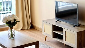 Moderni olohuone, jossa on televisio puisen lipaston päällä, valkoisia kukkia maljakossa pöydällä ja näkymä ulos ikkunasta.