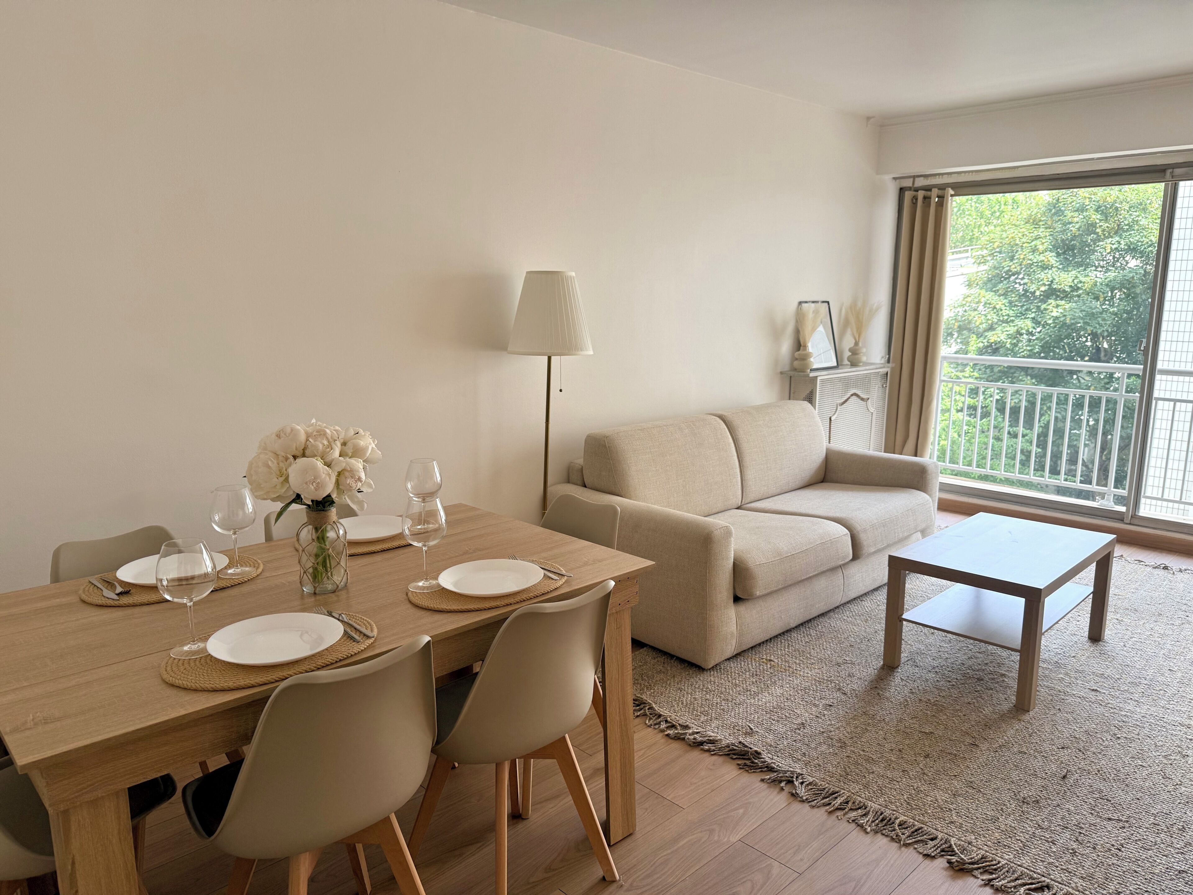 A modern living room with a dining table set for four, a sofa, a coffee table, and a large window with a view of greenery.
