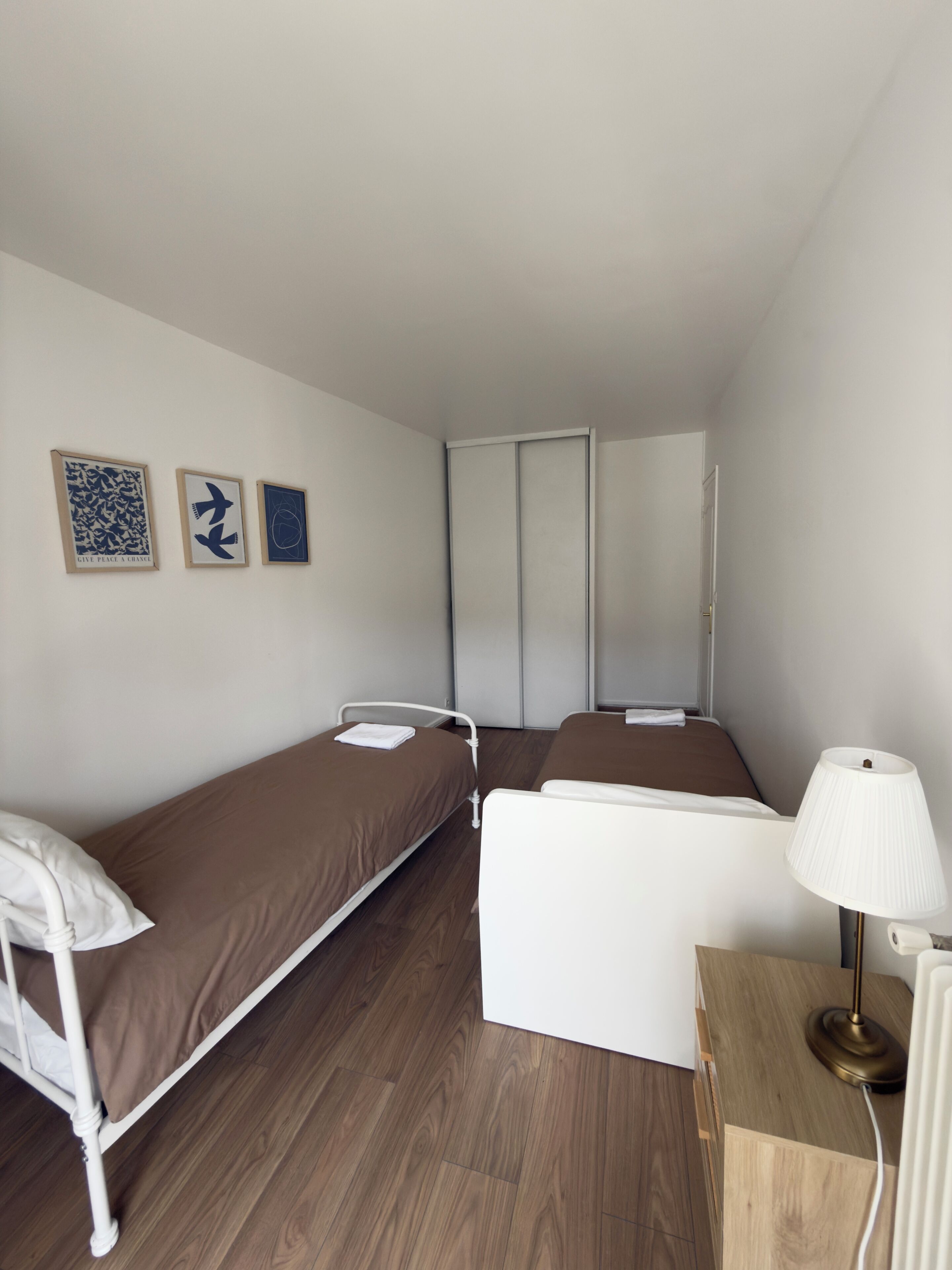 Une petite chambre d’hôtel composée d’une seule pièce, avec deux lits simples, une table de chevet avec une lampe, un plancher en bois et trois œuvres d’art encadrées accrochées au mur.