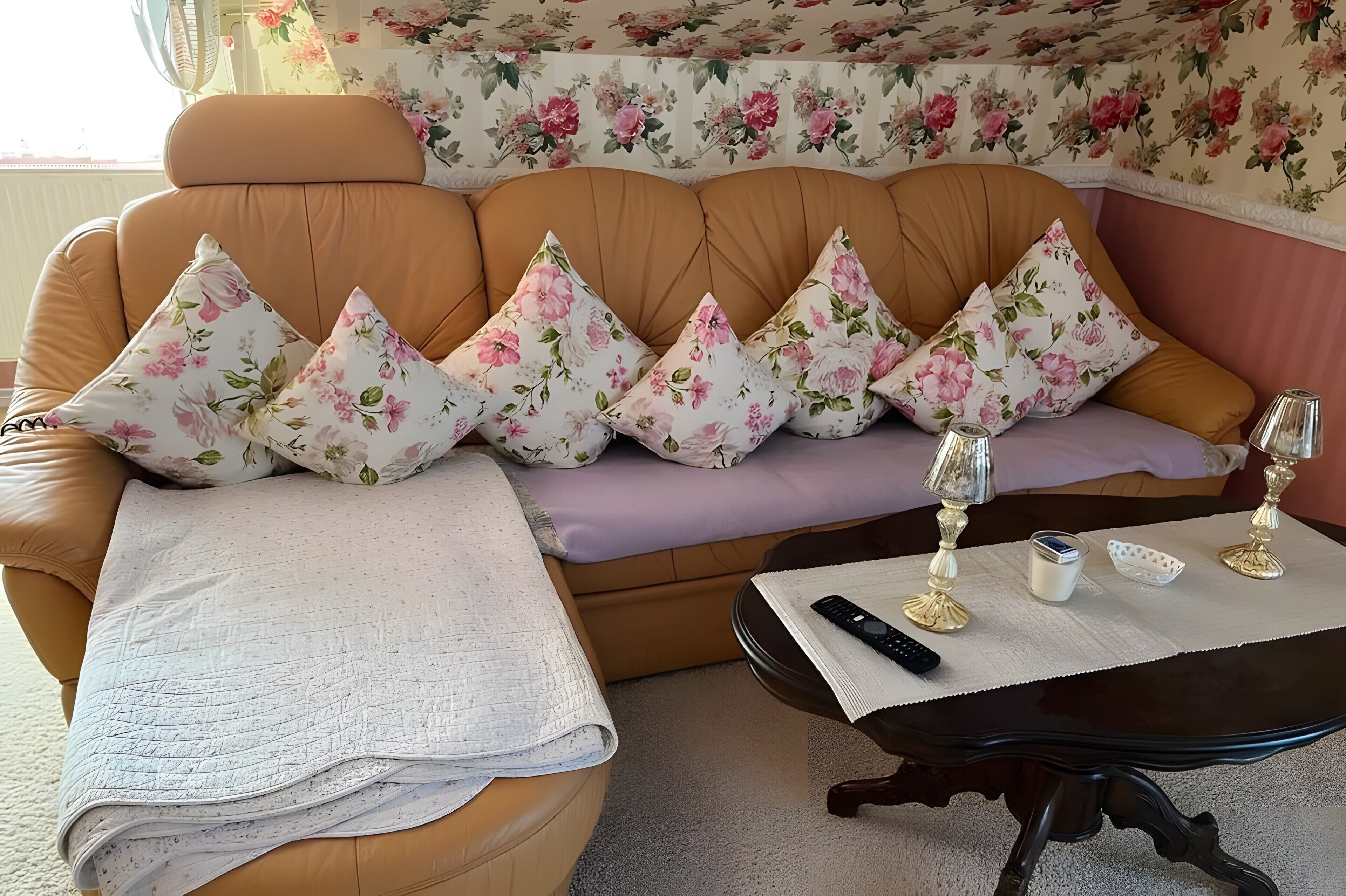 Un soggiorno con un divano marrone, cuscini a motivi floreali, una coperta bianca, un tavolino con un telecomando, un bicchiere e una tazza.