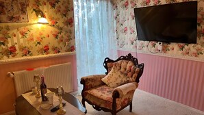 A room with a floral wallpaper, a television mounted on the wall, a wooden chair, a table with a lamp, and a radiator. - Vacation Apartment in a Country House in a Quiet Setting in the Elm-Lappwald Recreation Area (Frellstedt)