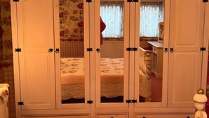 A room with a large wardrobe, a bed with floral bedding, a window with curtains, and a mirror. - Vacation Apartment in a Country House in a Quiet Setting in the Elm-Lappwald Recreation Area (Frellstedt)