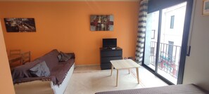 Ein Zimmer mit einer Couch, einem kleinen Tisch, einem Fernseher auf einem Schrank und einem Balkon mit Aussicht.
