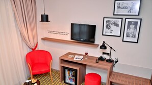 A compact hotel room with a TV, a small fridge, a red chair, a wooden desk, and a patterned floor. - Capitolina (Cluj-Napoca)