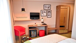 A hotel room with a bed, a desk with a TV, a red chair, a wardrobe, and a small fridge. - Capitolina (Cluj-Napoca)
