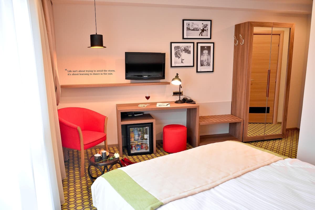 Habitación de hotel con cama, escritorio con televisor, silla roja, armario y refrigerador pequeño.