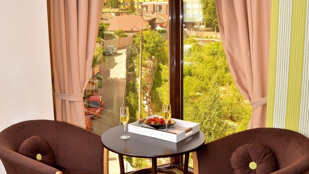 A room with two brown armchairs, a small round table with a glass of wine, and a view of a cityscape through large windows. - Capitolina (Cluj-Napoca)