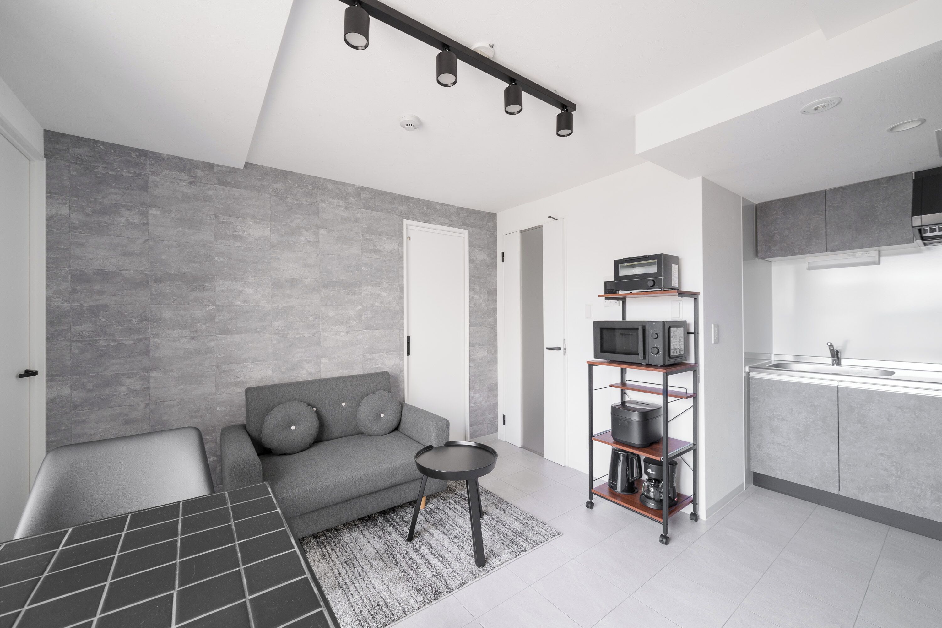 Un espacio habitable moderno con un sofá gris, una pequeña mesa redonda, una zona de cocina con armarios grises y un microondas, y un piso de baldosas grises.
