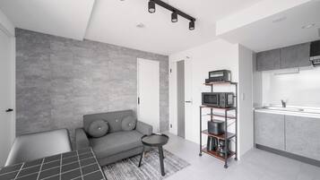Un espacio habitable moderno con un sofá gris, una pequeña mesa redonda, una zona de cocina con armarios grises y un microondas, y un piso de baldosas grises.
