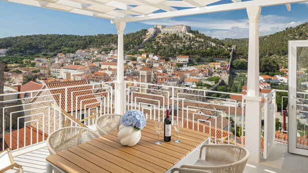 Terraço com mesa de madeira, cadeiras de vime branco e vista para um castelo histórico no alto da colina.