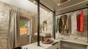 Um quarto com parede de pedra, uma janela com cortinas, uma mesa com chapéu e um cabideiro com várias peças de roupa.