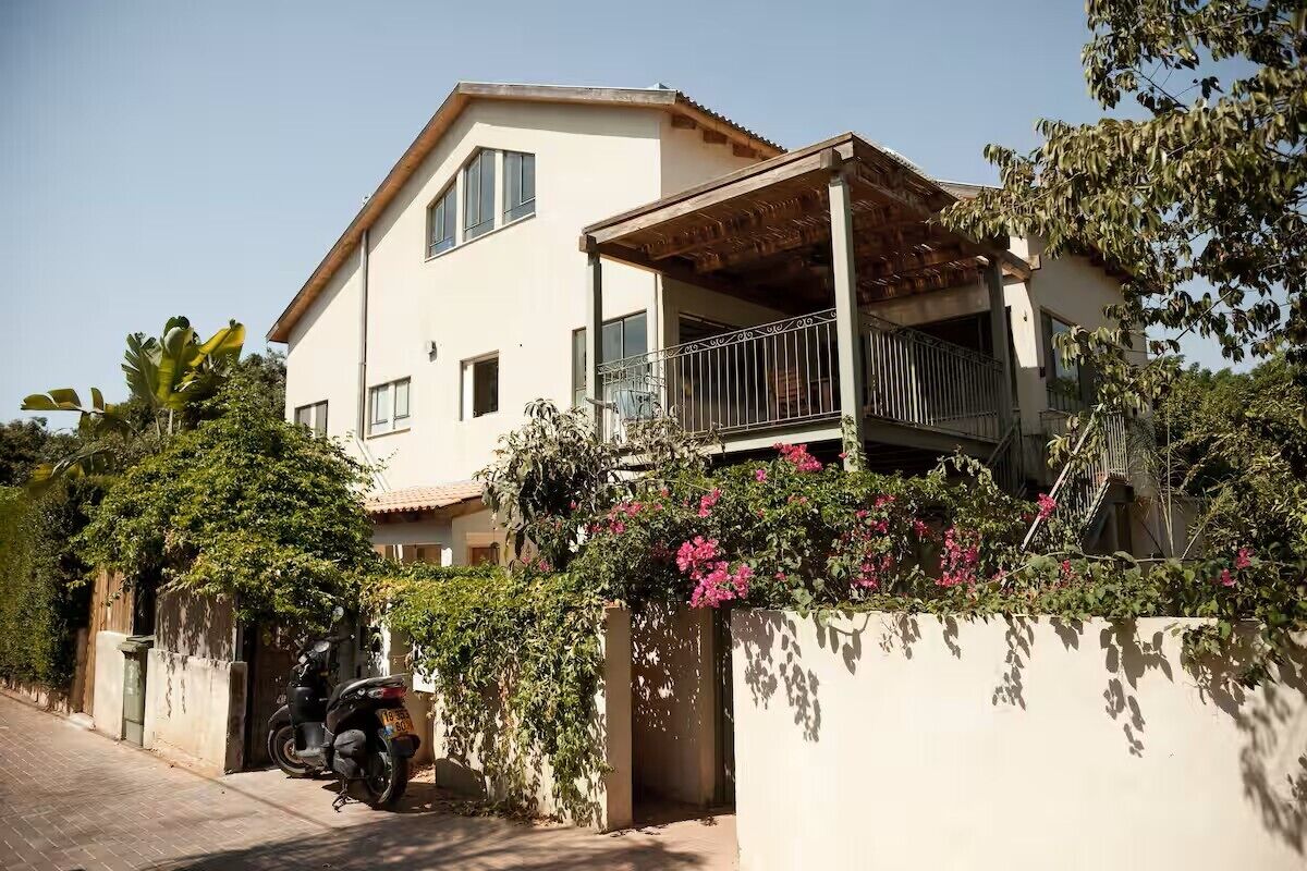 A two-story house with a balcony, a motorcycle parked in front, and a wall covered with greenery and flowers.