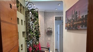 Un couloir avec une porte en bois, un paravent décoratif en métal agrémenté de plantes, une chaise rouge, une table blanche et un tableau représentant un paysage urbain accroché au mur.