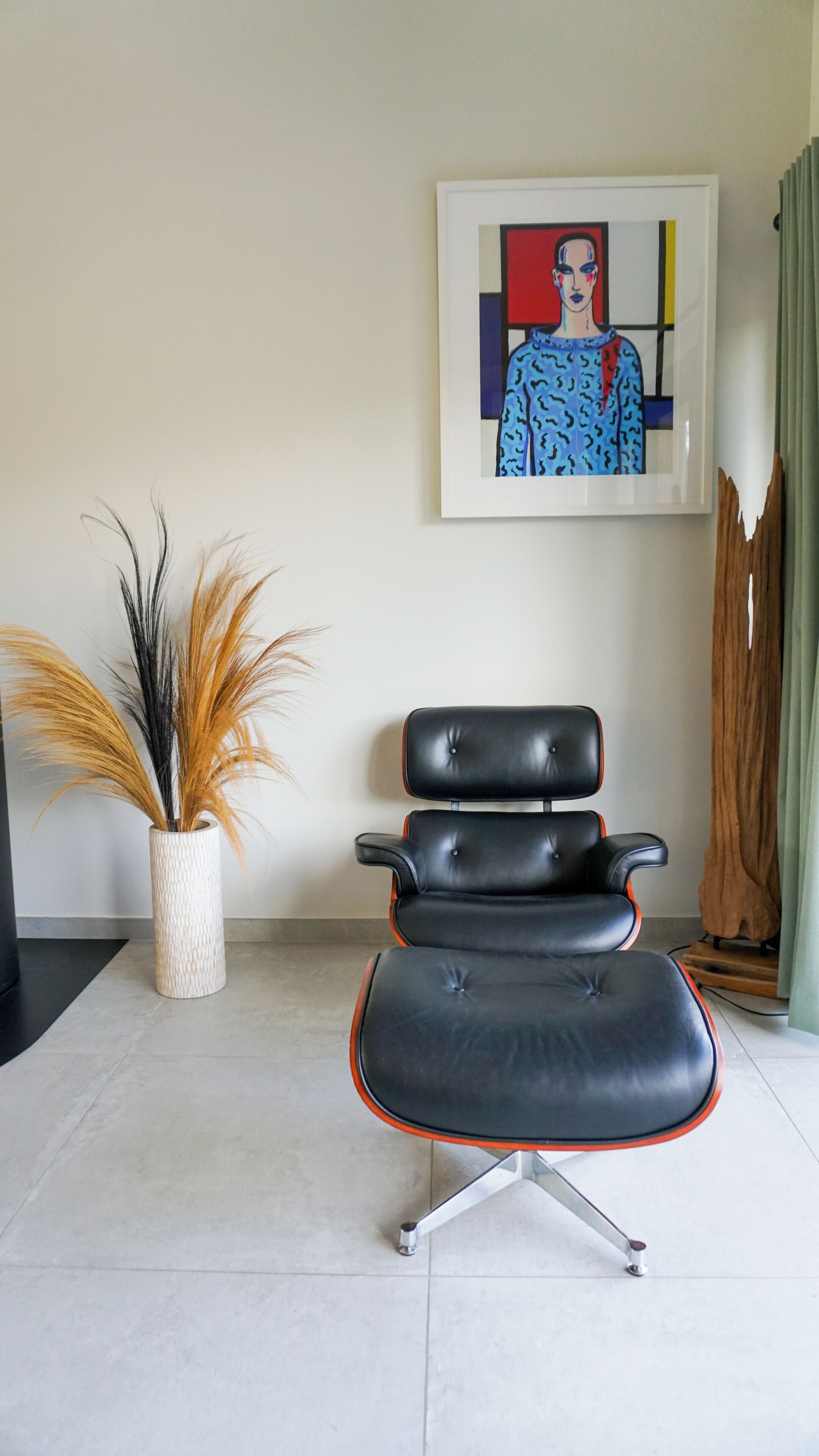 Een moderne stoel met een hoge rugleuning en armleuningen, een witte vaas met gedroogde planten, en een abstract schilderij in een lijst aan de muur.