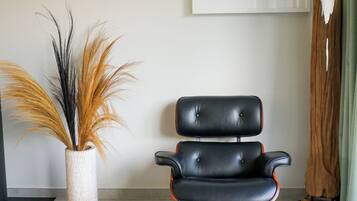 Een moderne stoel met een hoge rugleuning en armleuningen, een witte vaas met gedroogde planten, en een abstract schilderij in een lijst aan de muur.