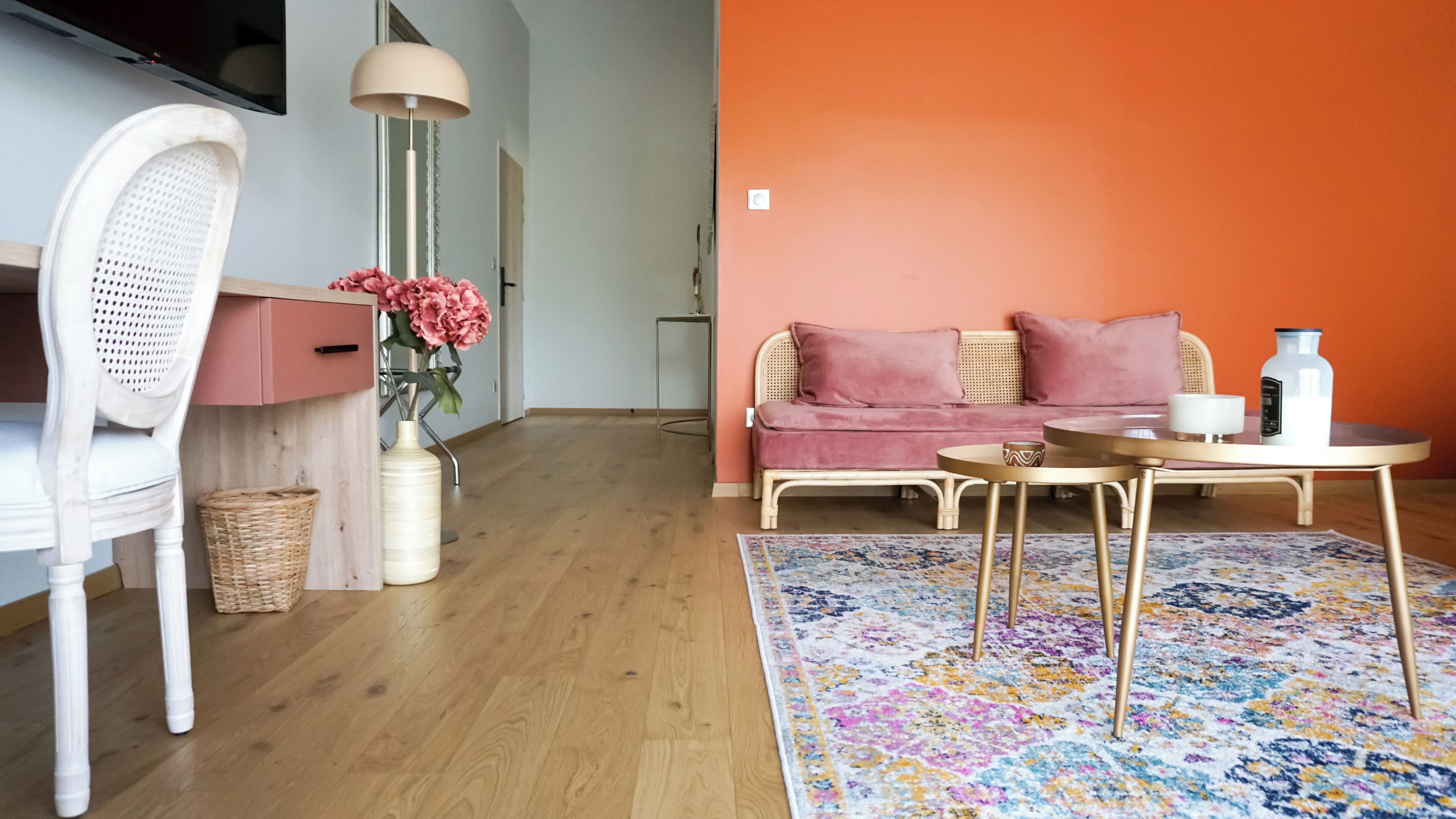 Ett rum med en vit stol, en soffa med rosa kuddar, ett litet runt bord och en färgglad matta.