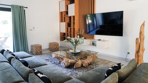Un salon moderne avec une table basse en verre, un tĂ©lĂ©viseur Ă Ă©cran plat, une bibliothĂšque en bois et une grande fenĂȘtre avec des rideaux.