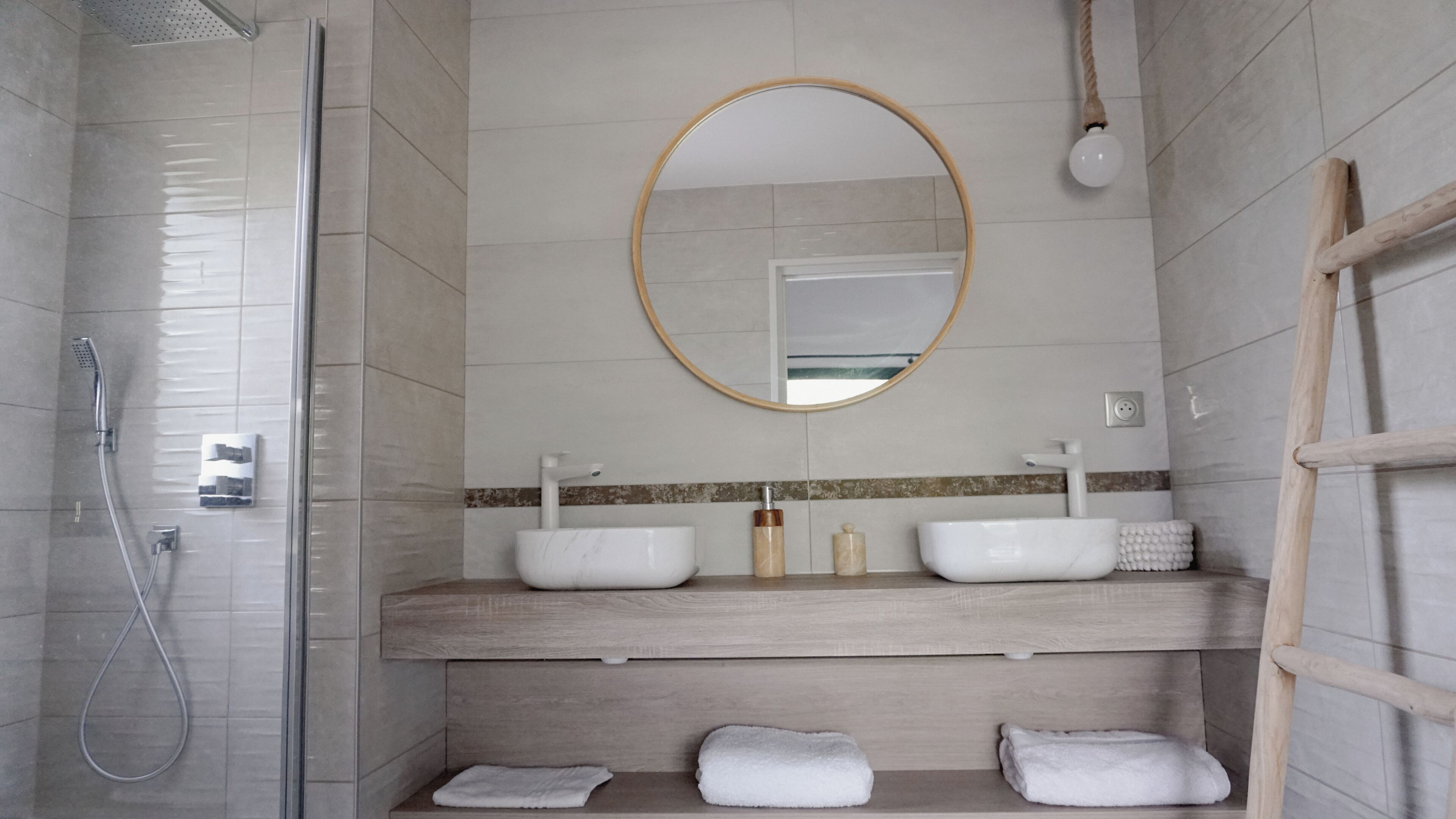 A modern bathroom with a glass shower enclosure, a wooden vanity with two sinks, a round mirror, and a towel rack.