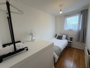 A bedroom with a bed, a bedside table with a lamp, a window with curtains, a standing fan, and a radiator. - Apartment Sleeps 6 - Wifi & Free Parking (Mitcham)