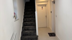 Een gang met een tapijtvloer, een wenteltrap, een radiator, een deur met een bordje en een lamp aan het plafond.