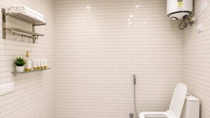 A bathroom with white subway tiles, a toilet, a shower, and shelves with toiletries. - Govind Residency (Greater Noida)