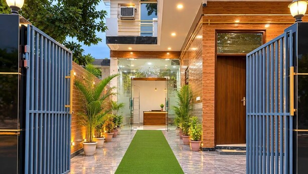 A modern building entrance with a reception desk, potted plants, and a green artificial turf path. - Govind Residency (Greater Noida)