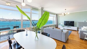 A modern living room with a large window, a dining area, and a sofa. - Azure Deluxe • Central Bosphorus View Flat (Beyoglu)
