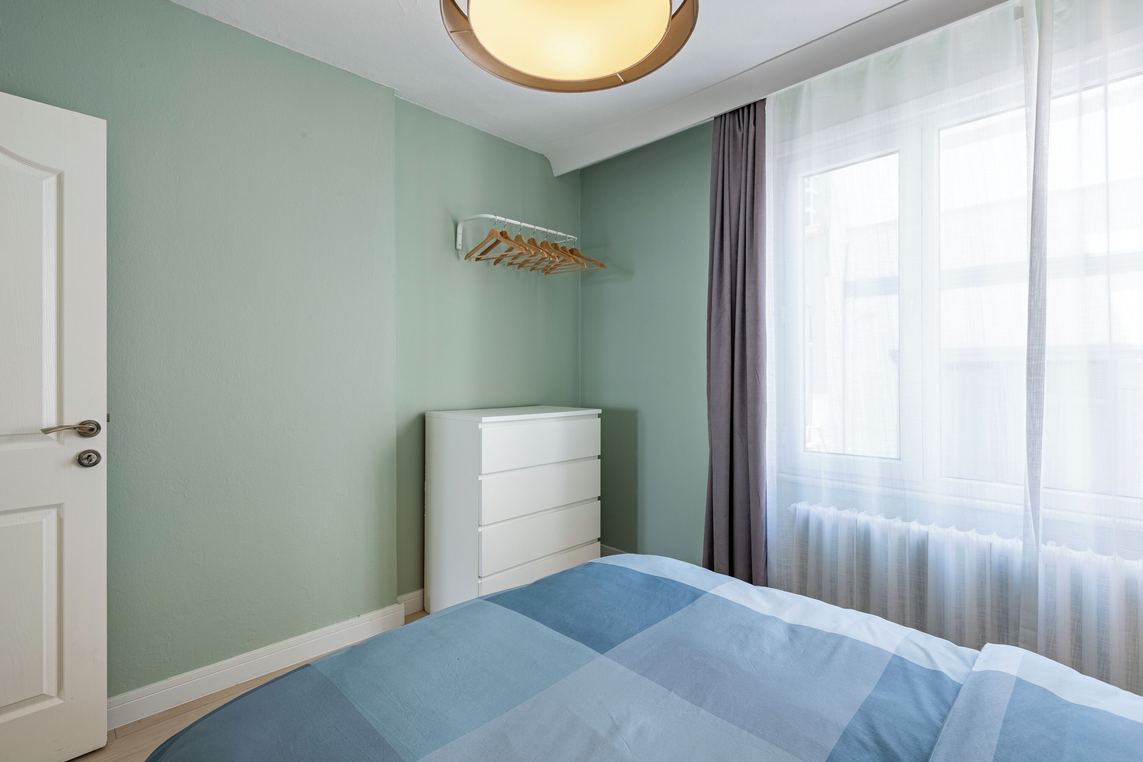 Um quarto com uma cama, cômoda, porta e janela com cortinas.
