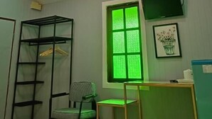 Una habitación con literas, un escritorio, una silla, una ventana iluminada en verde, un televisor en la pared y una lámpara en el techo.