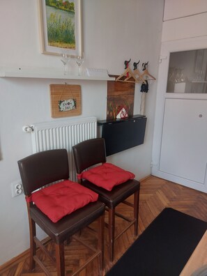 A room with two wooden chairs with red cushions, a radiator, a wooden sign with text, a shelf with decorative items, and a window with a view of greenery. - Különálló apartman,légkondicionált,saját fürdőszobával. (Budapest)