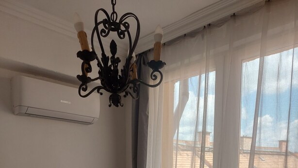 Une chambre avec un lit, un bureau avec une lampe, une chaise, une fenêtre avec des rideaux légers et un lustre.