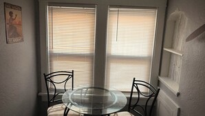 Un área de comedor con una mesa redonda de vidrio y cuatro sillas, dos ventanas con persianas, y un cartel colgado en la pared.