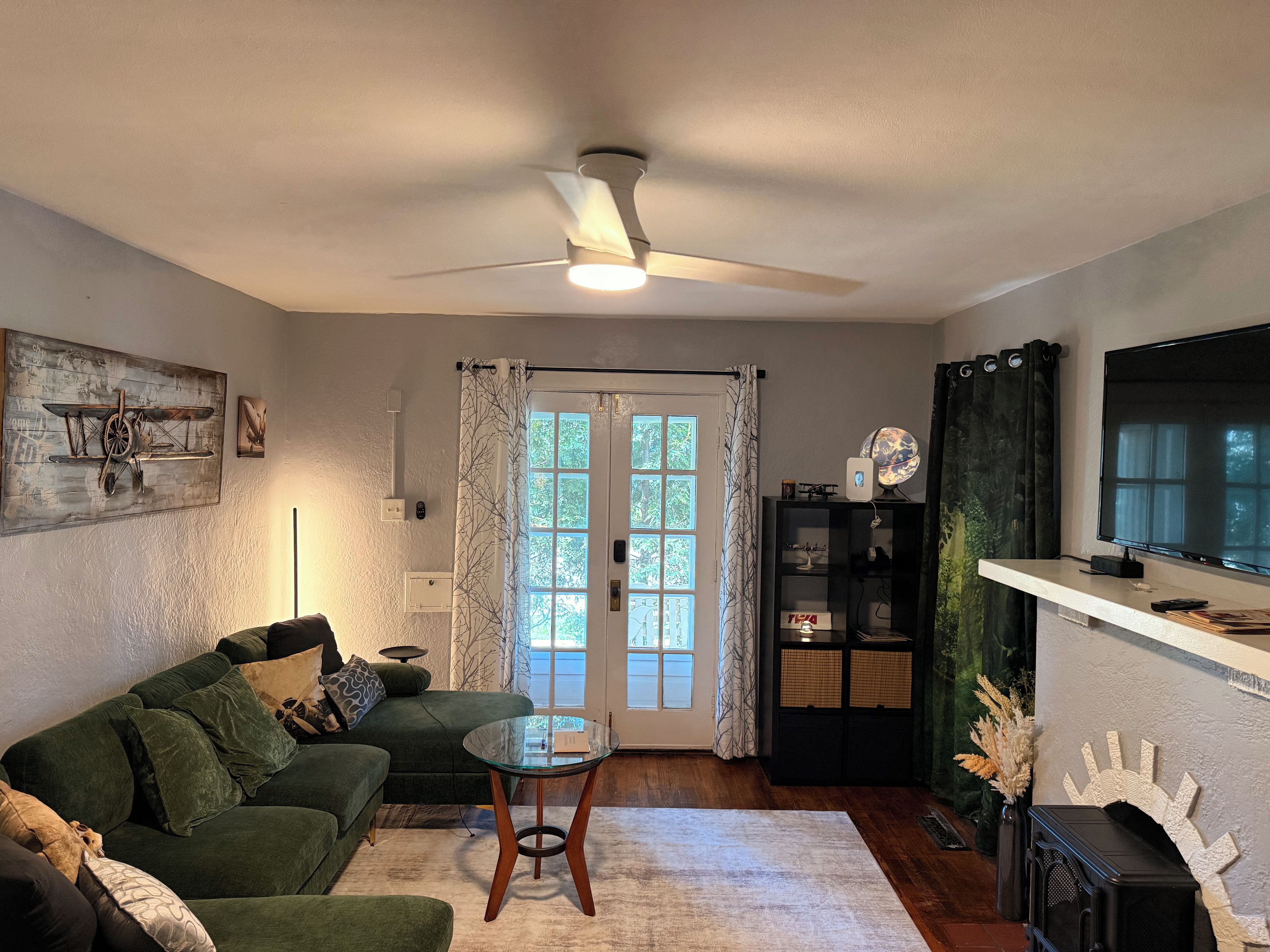 Un salon avec un canapé vert, une table basse en verre, un ventilateur de plafond, un téléviseur et une bibliothèque.