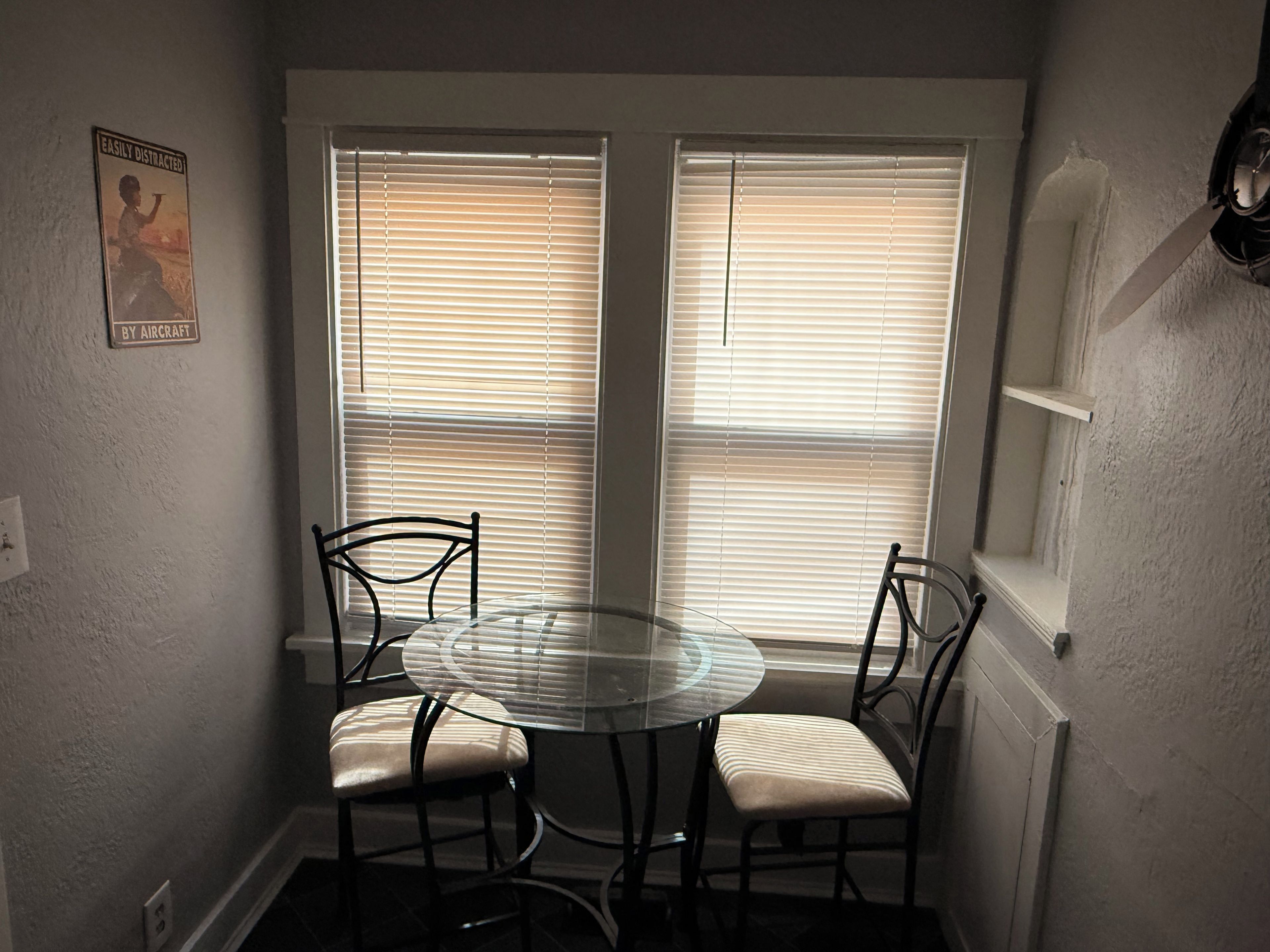 Espaço de refeições com mesa de vidro redonda e quatro cadeiras, duas janelas com persianas e luminária fixada na parede.