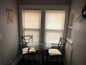 A dining area with a round glass table and four chairs, two windows with blinds, and a wall-mounted light fixture.
