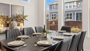 Comedor con una mesa larga para seis personas, ventanas grandes y vista a una casa.
