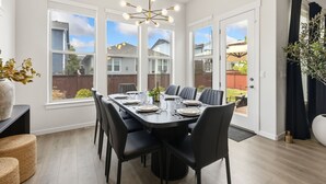 Comedor con una gran mesa dispuesta para seis personas, una lámpara moderna y ventanas amplias que dan al jardín.